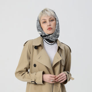 a black and white silk scarf with birds, flowers and trees prints worn by a women in a classic beige coat, wrapped on her head as a head scarf