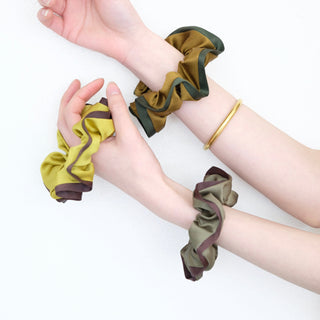 three silk scrunchies in different colours holding by a pair of woman's hands, one is mustard with brown edge, one is copper with dark green edge, one is olive green with brown edge