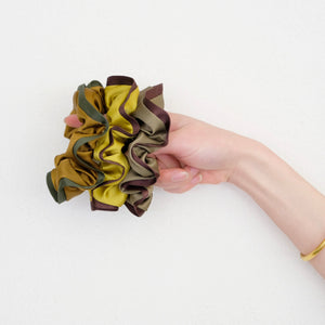 three silk scrunchies in different colours holding by a woman's hand, one is mustard with brown edge, one is copper with dark green edge, one is olive green with brown edge