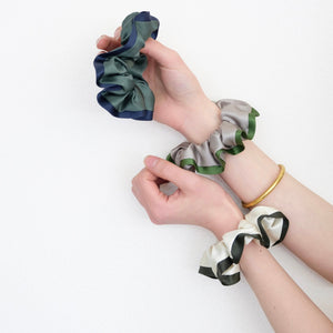 three silk scrunchies in different colours holding by a pair of woman's hands, one is khaki with green edge, one is creamy white with dark green edge, one is green with blue edge