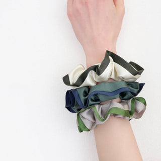 three silk scrunchies in different colours worn on a woman's wrist, one is khaki with green edge, one is creamy white with dark green edge, one is green with blue edge