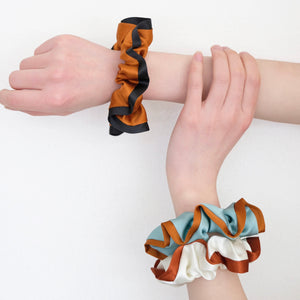 three silk scrunchies in different colours holding by a pair of woman's hands, one is burnt orange with black edge, one is mint blue with orange edge, one is off white with rus red edge