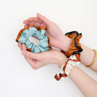 three silk scrunchies in different colours holding by a pair of woman's hands, one is burnt orange with black edge, one is mint blue with orange edge, one is off white with rus red edge