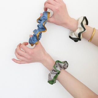 three silk scrunchies in different colours holding by a pair of woman's hands, one is khaki with green edge, one is creamy white with dark green edge, one is Egyptian blue with bronze edge