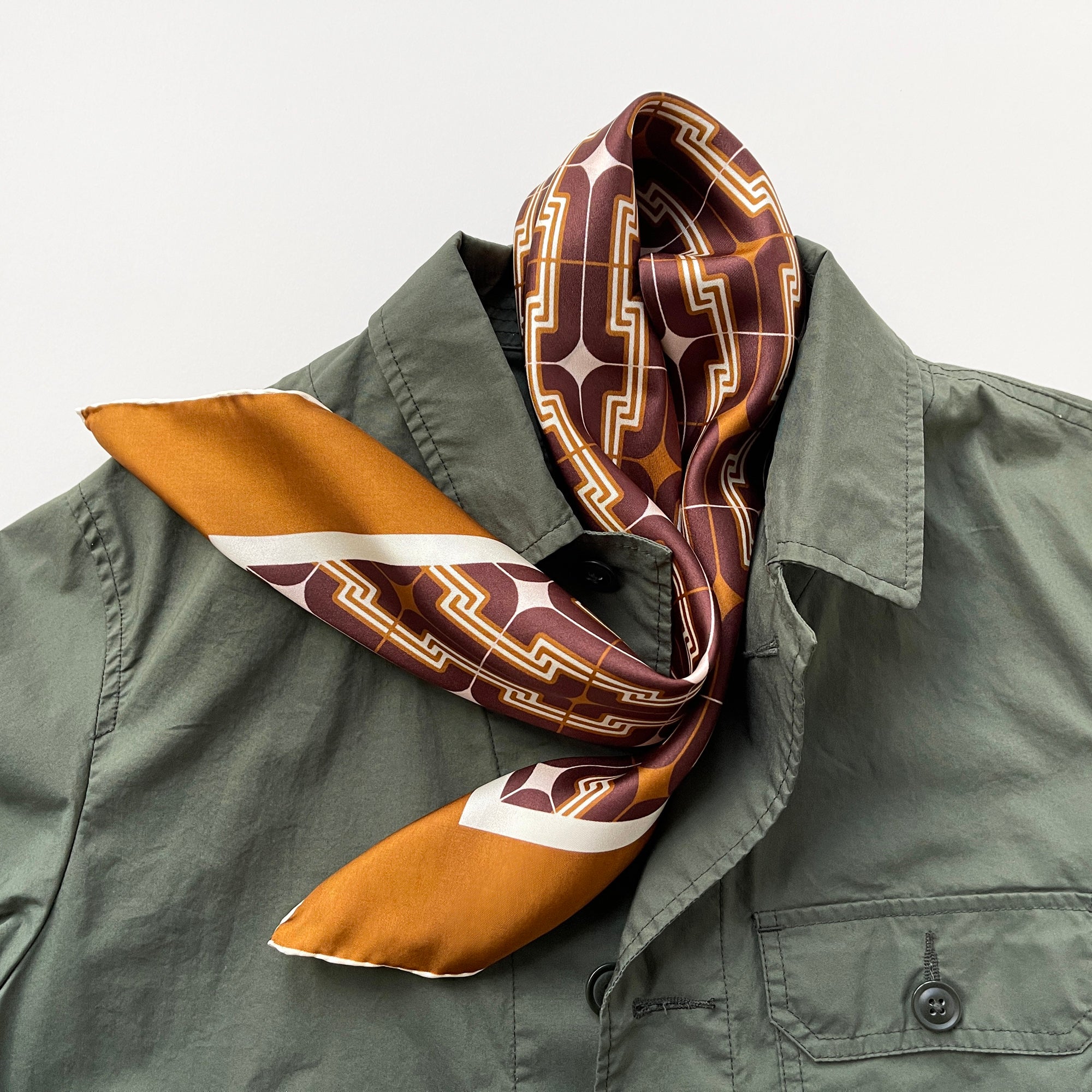 a luxury silk scarf featuring vintage charm 70s style patten in burgundy, tan and beige palette with hand-rolled hems, paired with a army green men's jacket