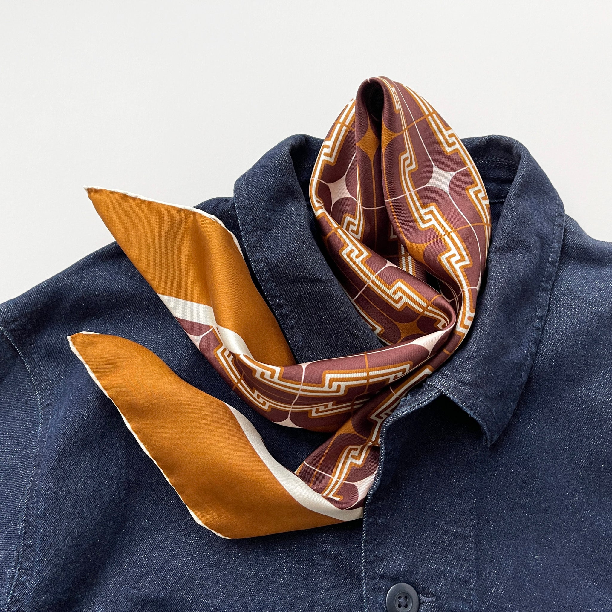 a luxury silk scarf featuring retro charm 70s style patten in burgundy, tan and beige palette with hand-rolled hems, paired with a denim jacket