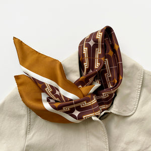 a luxury silk scarf featuring vintage charm 70s style patten in burgundy, tan and beige palette with hand-rolled hems, paired with a beige coat