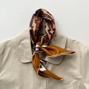 a luxury silk scarf featuring 70s style patten in burgundy, tan and beige palette with hand-rolled hems knotted as a neck scarf, paired with a beige coat