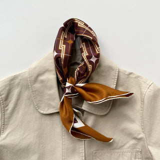 65x65cm brown silk scarf with a geometric pattern and hand-rolled edges, featuring a contrasting border. Retro-inspired unisex accessory, perfect for versatile styling around the neck, hair, or as an accent piece.