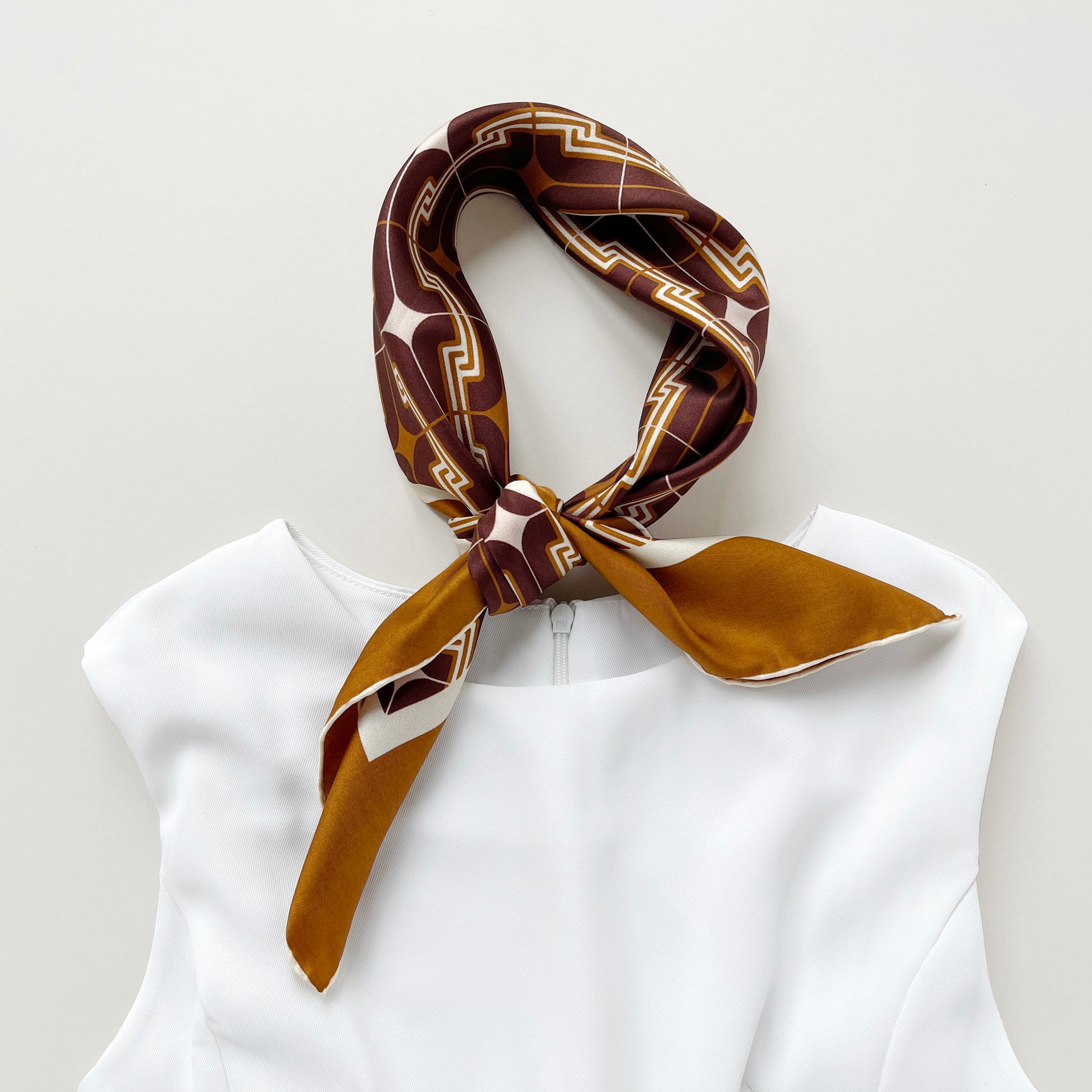 a luxury silk scarf featuring vintage charm 70s style patten in burgundy, tan and beige palette with hand-rolled hems knotted as a neckerchief, paired with a white sleeveless dress