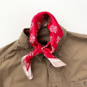 a red small silk scarf with beige edge featuring crown print, knotted as a neckerchief paired with a khaki men's shirt