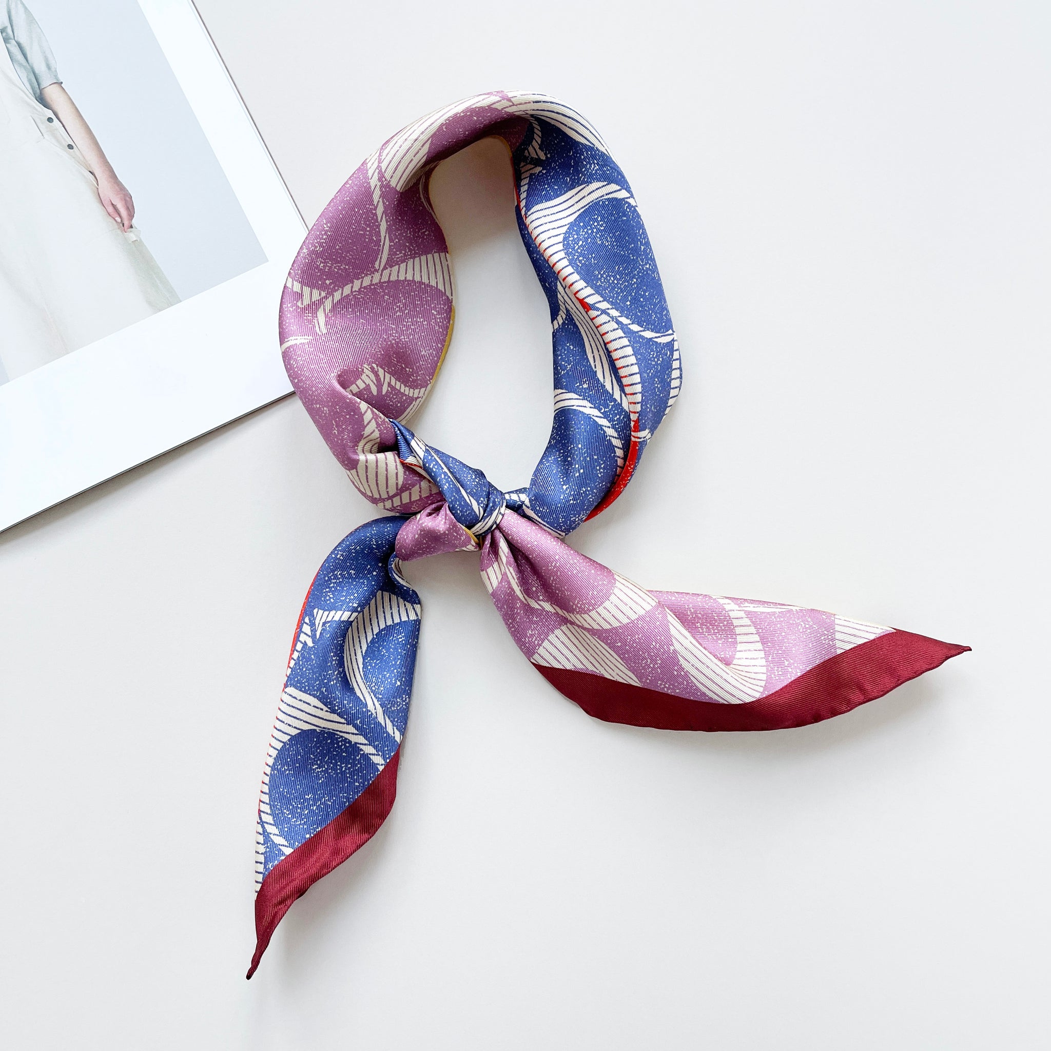 a luxury square silk scarf in lilac oink, blue, orange and yellow featuring burgundy hand-rolled edges and classic print, knotted as a neckerchief 