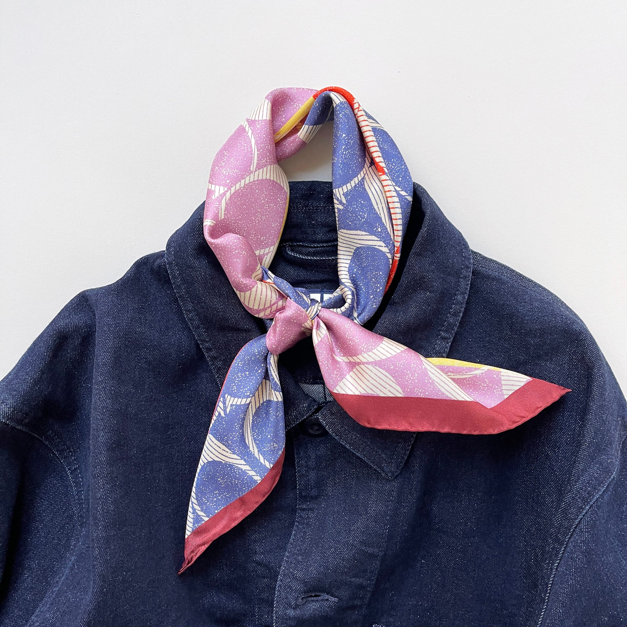 a luxury square silk scarf in lilac oink, blue, orange and yellow featuring burgundy hand-rolled edges and classic print, knotted as a neckerchief, paired with a unisex denim jacket