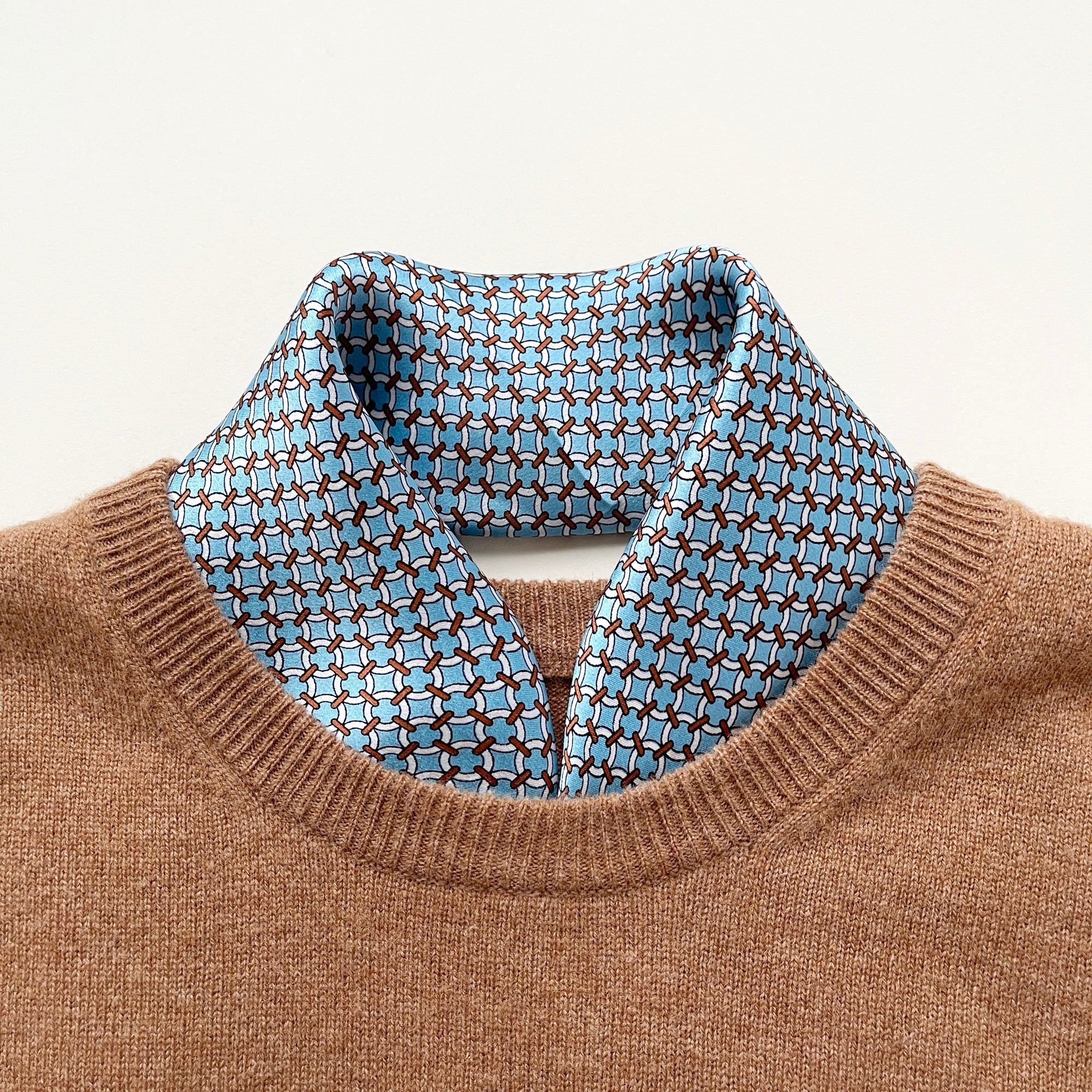 a small square silk scarf with brown edge featuring sky blue circle print, tucked in a camel men's sweater