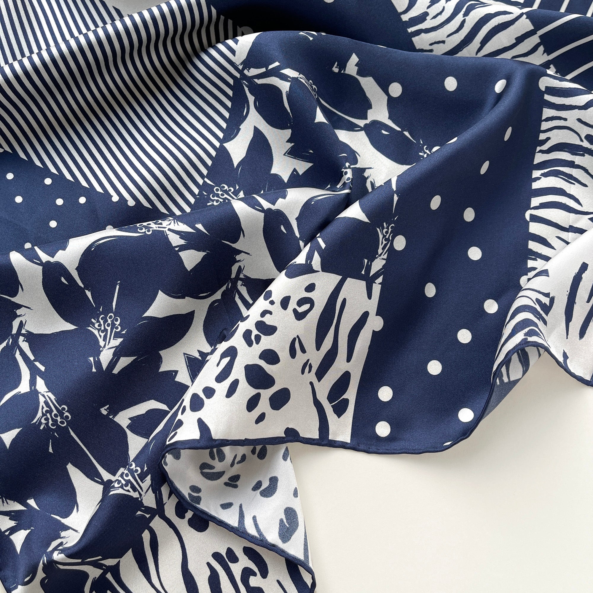 a navy blue and white square silk scarf featuring the combination of floral motifs, polka dots, and stripes prints with hand-rolled edges