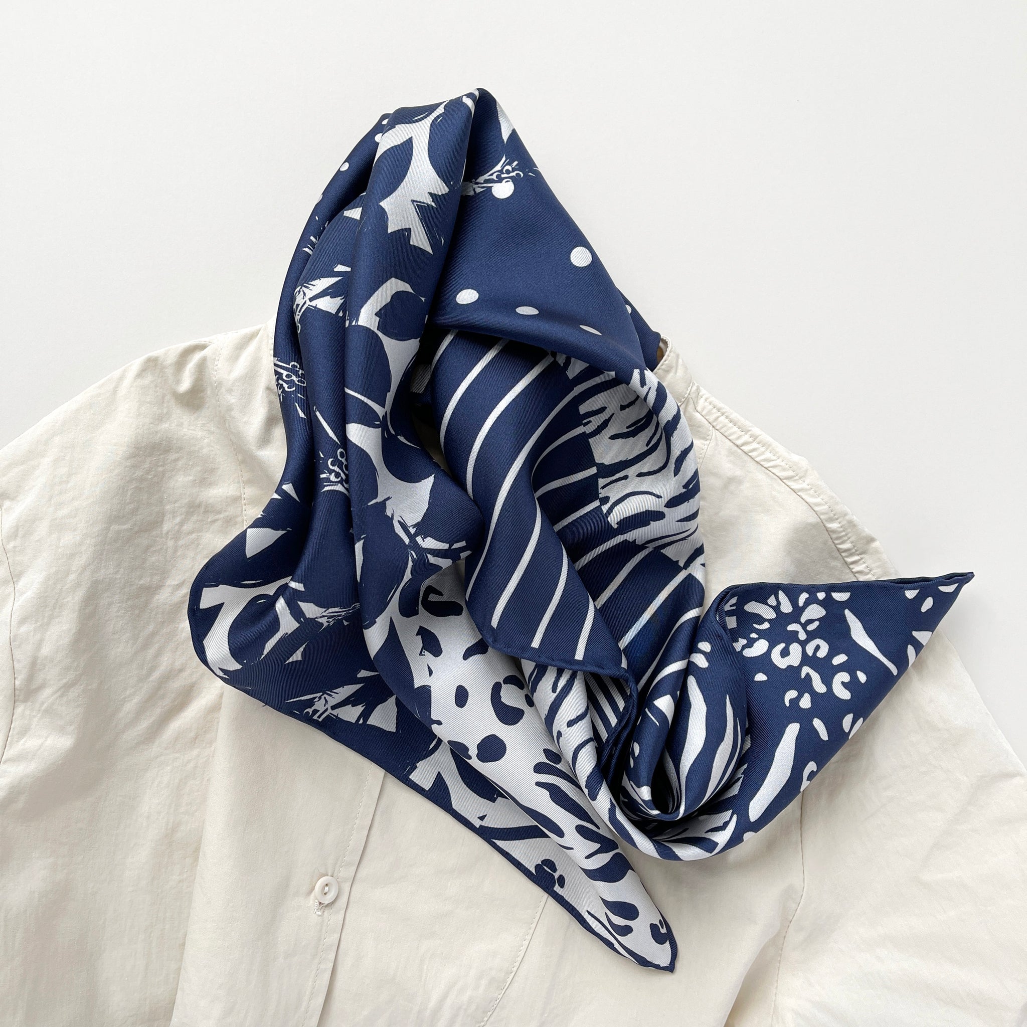 a navy blue and white square silk scarf featuring the combination of floral motifs, polka dots, and stripes prints with hand-rolled edges, paired with a light beige women's shirt