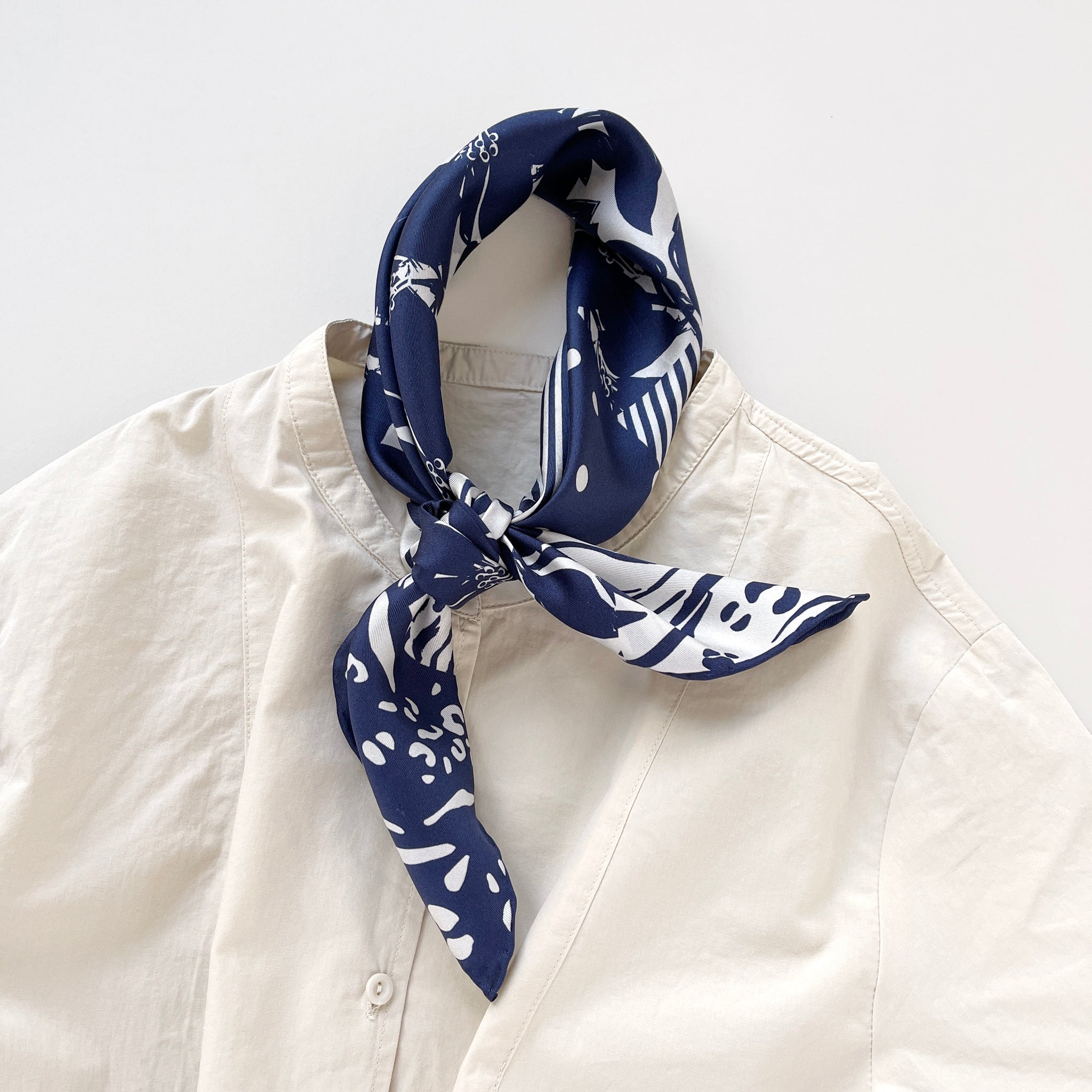 a white and blue square silk scarf featuring the combination of floral motifs, polka dots, and stripes prints with hand-rolled edges, knotted as a neckerchief, paired with a light beige women's shirt