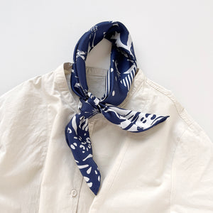 a white and blue square silk scarf featuring the combination of floral motifs, polka dots, and stripes prints with hand-rolled edges, knotted as a neckerchief, paired with a light beige women's shirt