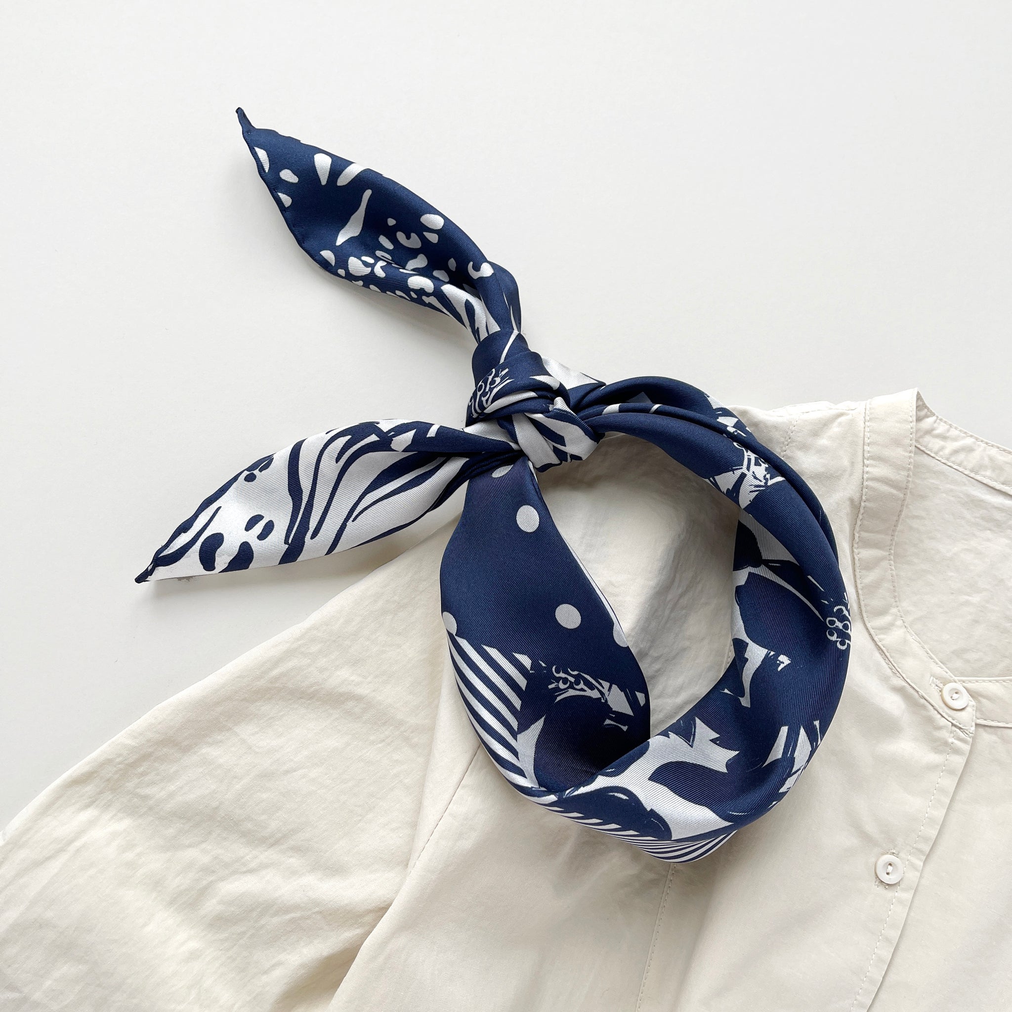 a navy blue and white square silk scarf featuring the combination of floral motifs, polka dots, and stripes prints with hand-rolled edges, tied as a headband, paired with a light beige women's shirt