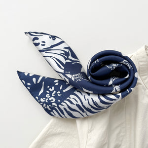 a navy blue and white square silk scarf featuring the combination of floral motifs, polka dots, and stripes prints with hand-rolled edges, paired with a light beige women's shirt