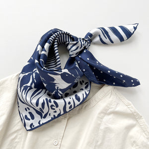 a navy blue and withe square silk scarf featuring the combination of floral motifs, polka dots, and stripes prints with hand-rolled edges, tied as a neck scarf, paired with a light beige women's shirt