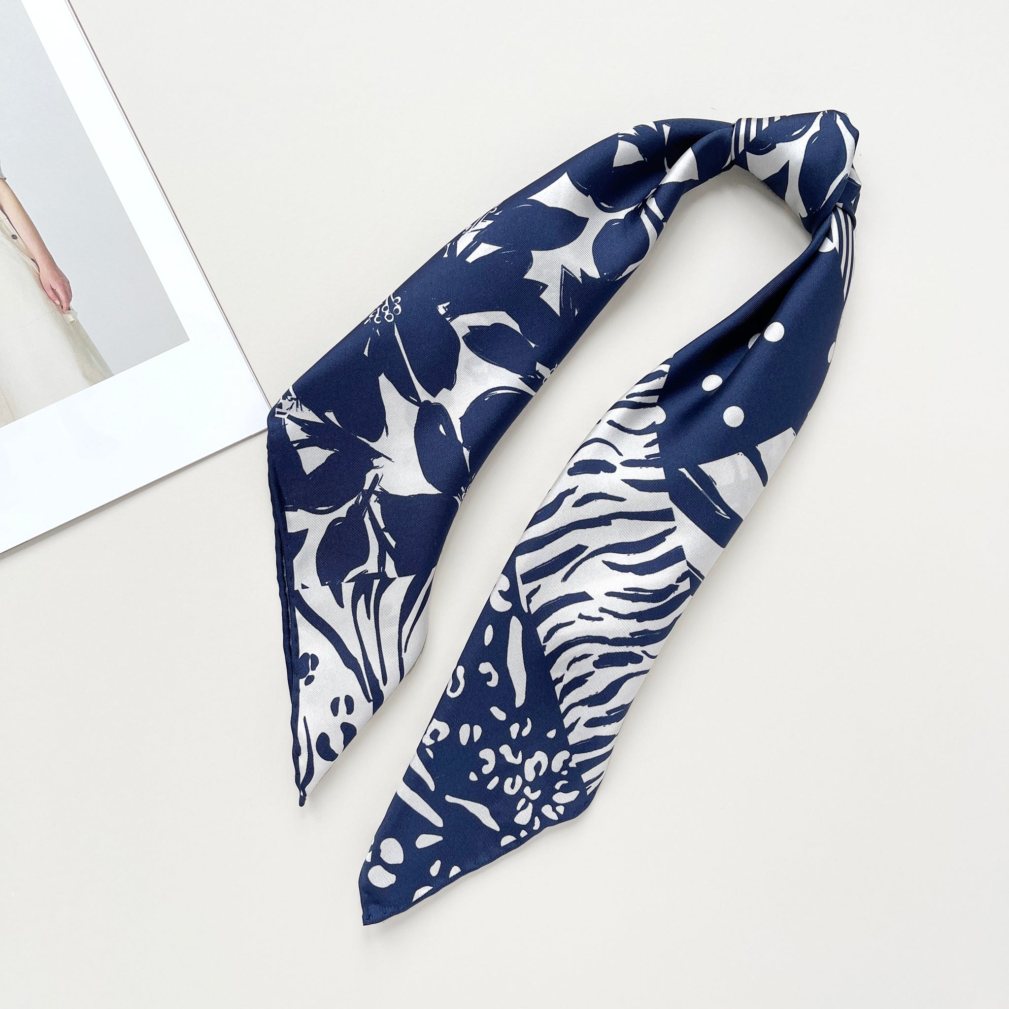 a navy blue and white square silk scarf featuring the combination of floral motifs, polka dots, and stripes prints with hand-rolled edges, tied as a ponytail 