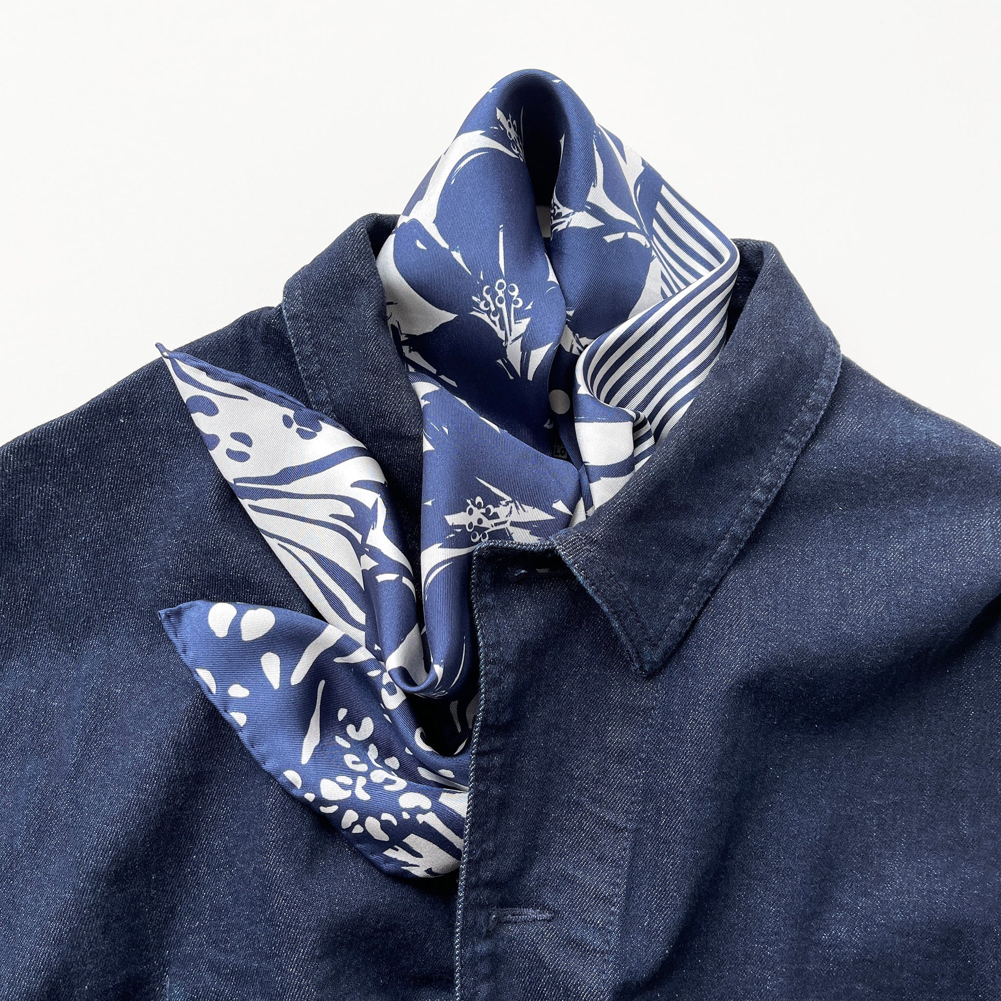 a navy blue and white square silk scarf featuring the combination of floral motifs, polka dots, and stripes prints with hand-rolled edges, paired with a denim jacket
