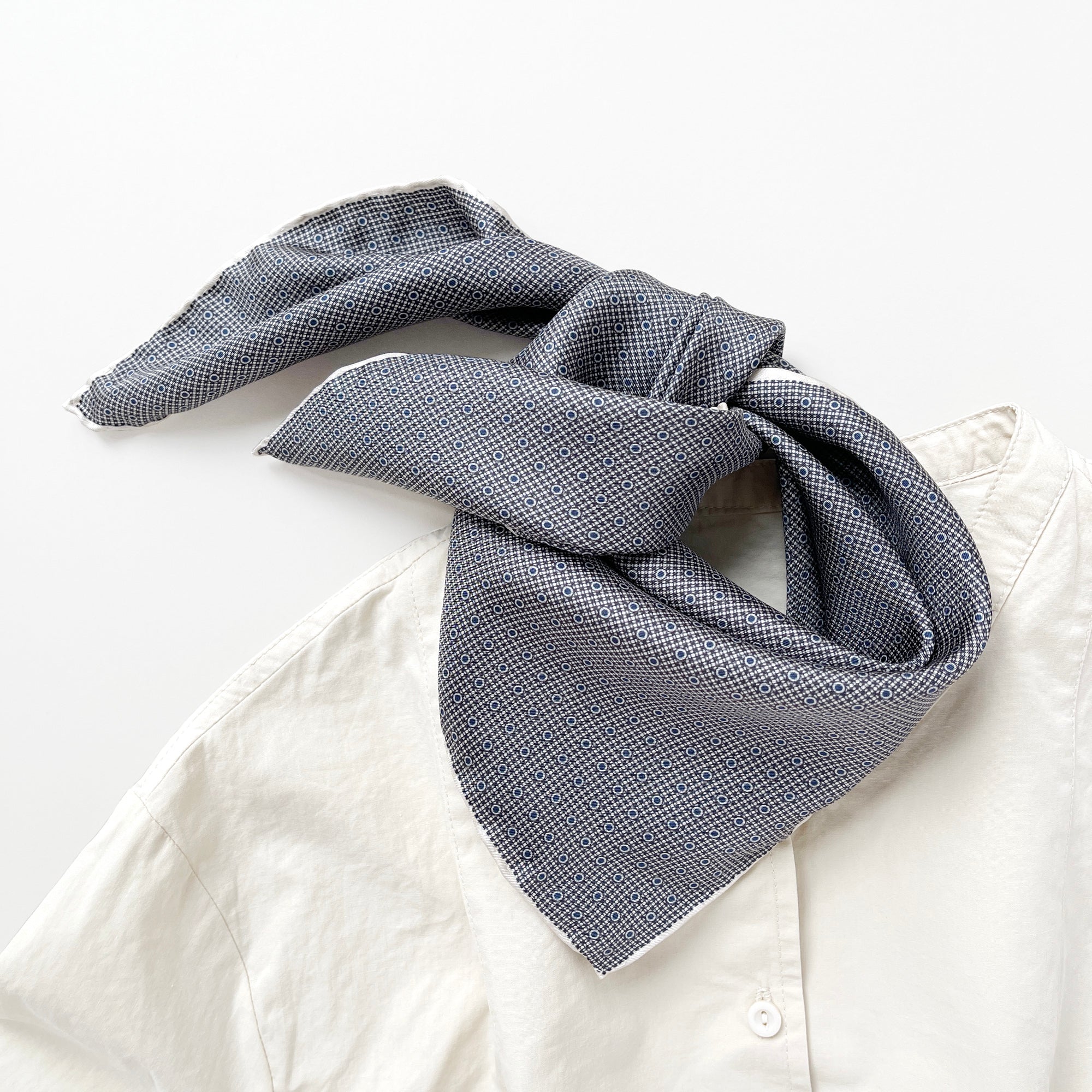 a classic silk scarf neckerchief featuring black and white checks and navy blue polka dots with white hand-rolled hems, tie around the neck as triangle fold, paired with a light beige shirt
