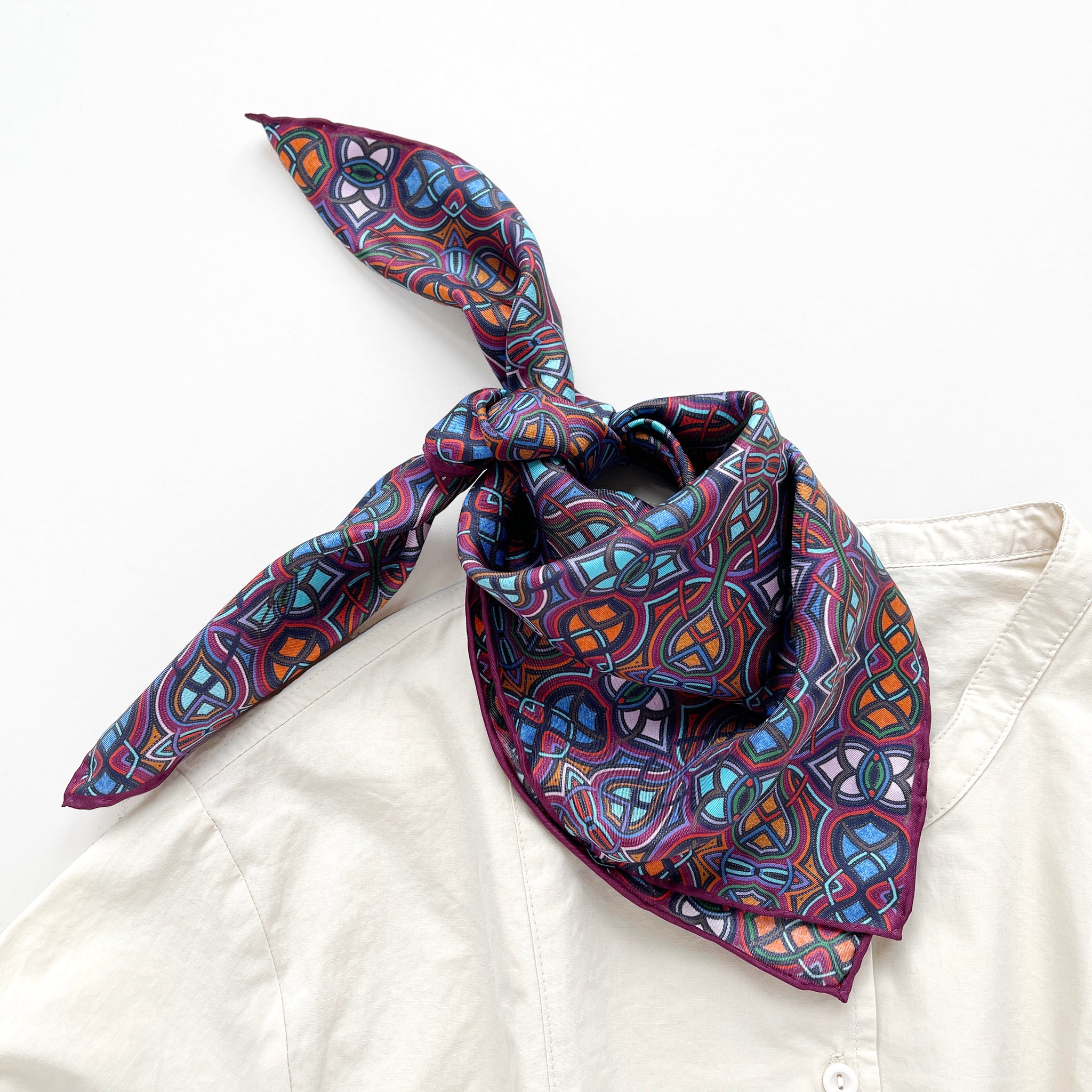 a small silk scarf neckerchief featuring an abstract intricate pattern in plum purple, blue and orange hues, tied as a bandana bib, paired with a light beige unisex shirt