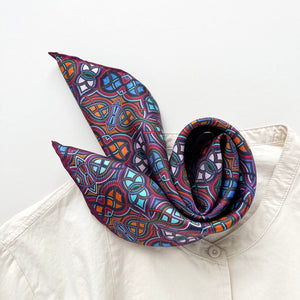a small silk scarf neckerchief featuring an abstract intricate pattern in plum purple, blue and orange hues paired with a light beige unisex shirt