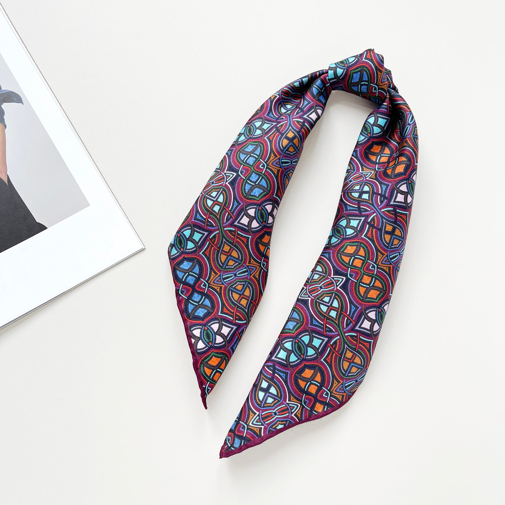 a silk neckerchief scarf featuring an abstract intricate pattern in plum purple, blue and orange hues, knotted as a ponytail