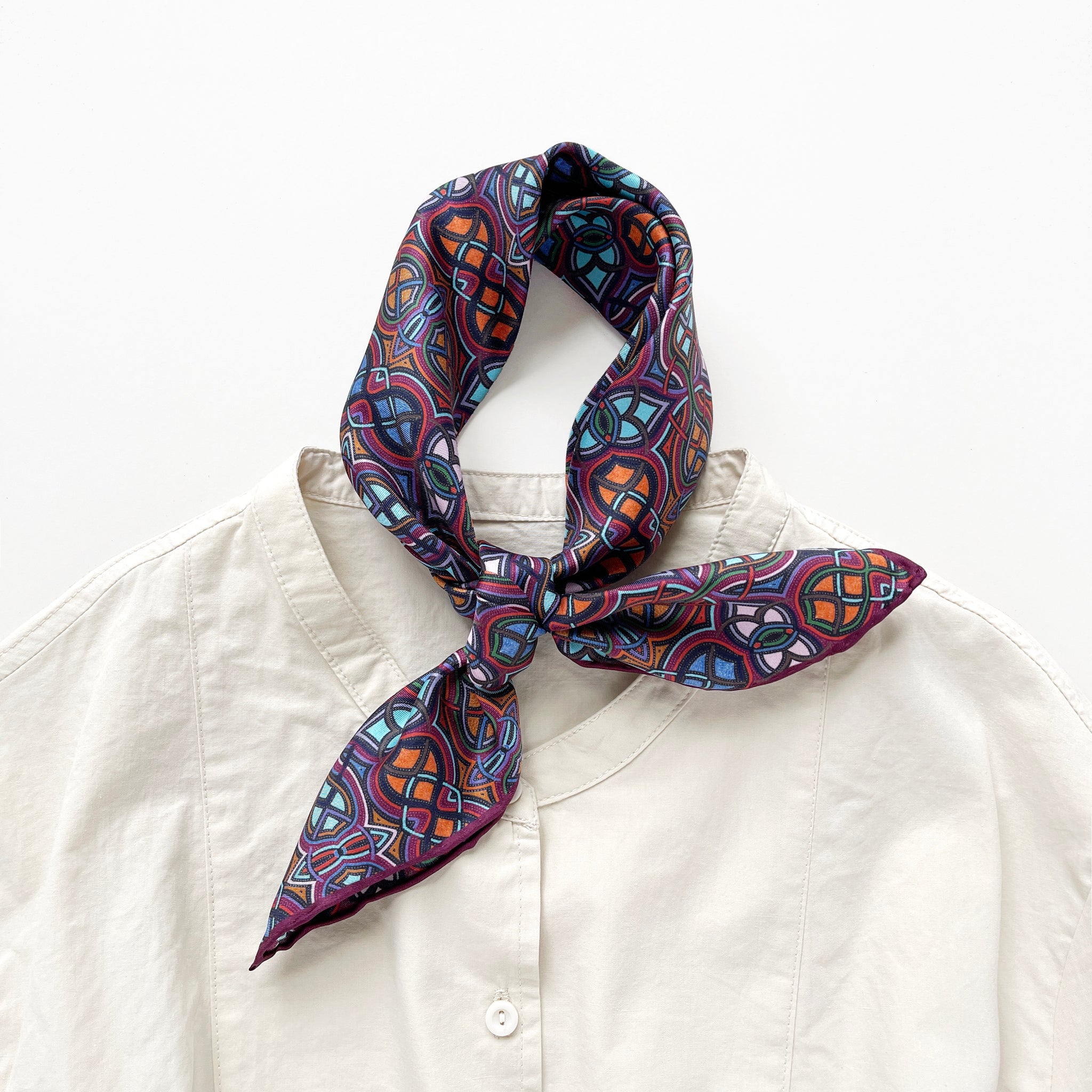 a small silk scarf neckerchief featuring an abstract intricate pattern in plum purple, blue and orange hues, knotted as a classic neckerchief, paired with a light beige unisex shirt