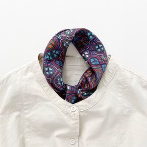 a small silk scarf neckerchief featuring an abstract intricate pattern in plum purple, blue and orange hues, knotted and tucked into a light beige unisex shirt