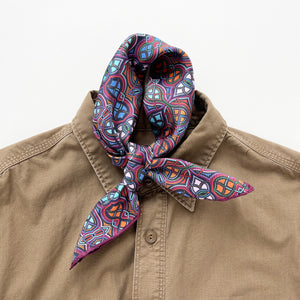 a silk men's neckerchief scarf featuring an abstract intricate pattern in plum purple, blue and orange, knotted as a classic neckerchief, paired with a men's khaki shirt