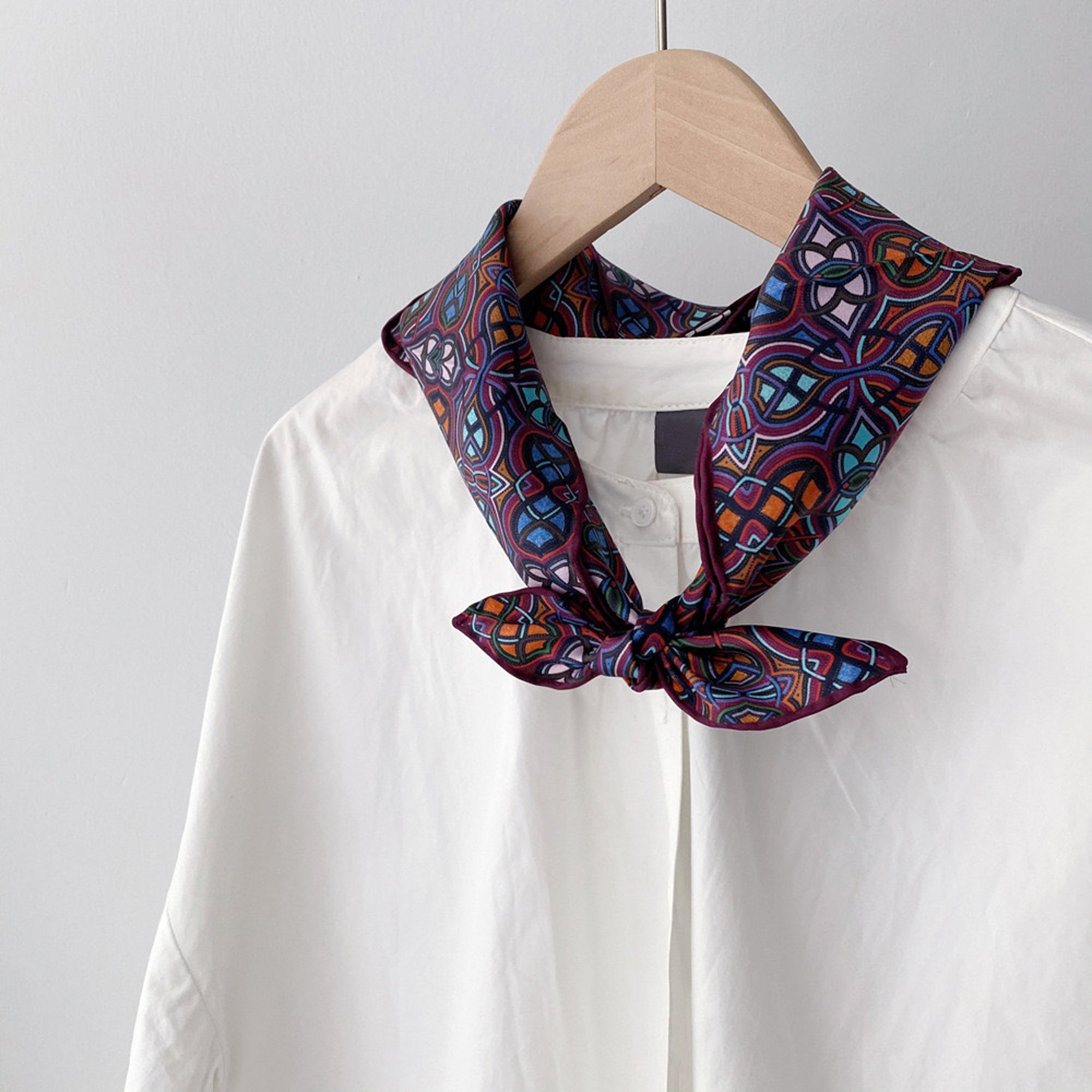 a small silk scarf featuring an abstract intricate pattern in plum purple, blue and orange, knotted as a neck scarf, paired with a women's white blouse