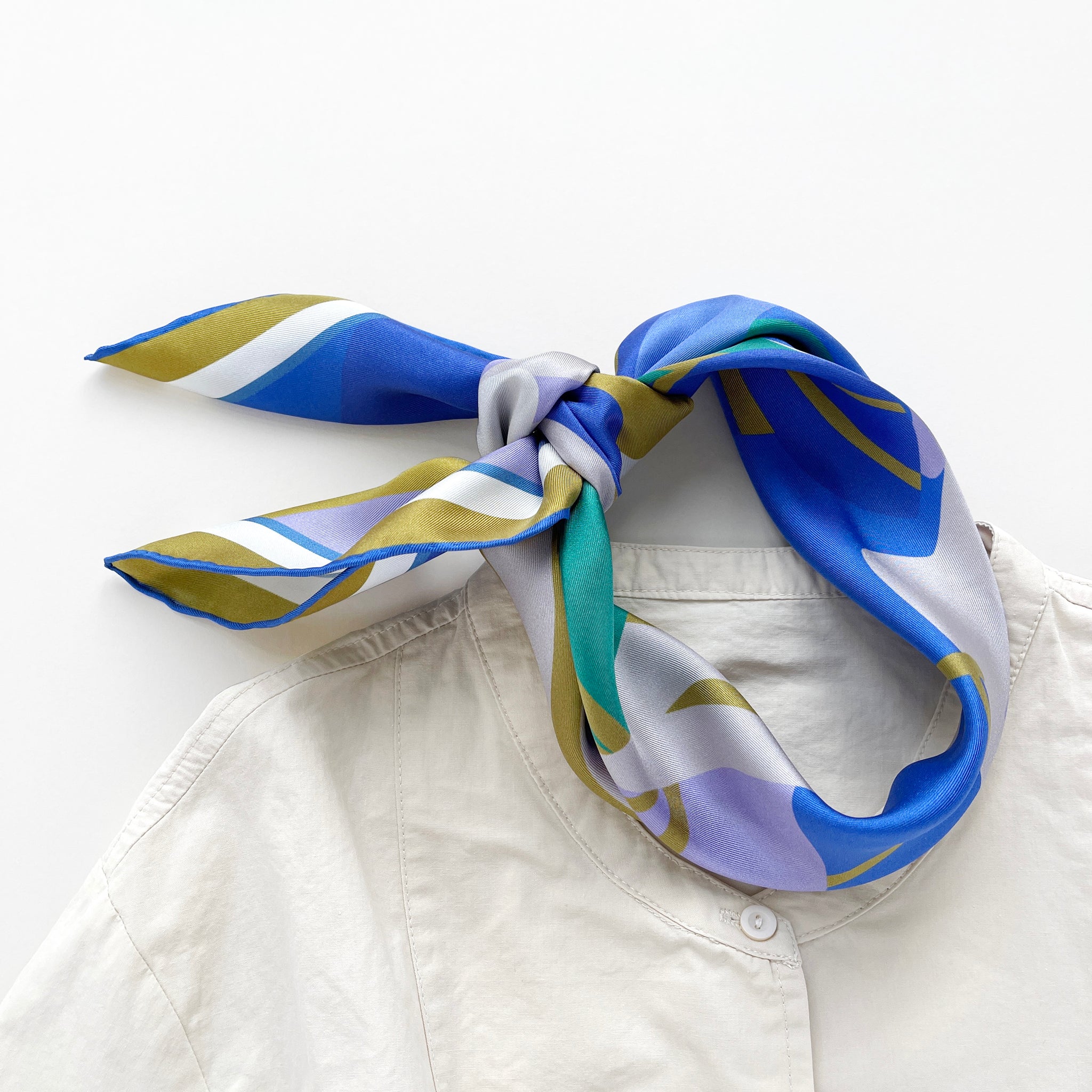 an abstract lotus print square silk scarf in blue, green, lilac and mustard yellow palette knotted as a neck scarf with hand-rolled hems paired with a light beige women's turtle neck shirt