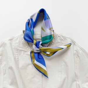 an abstract lotus print square silk scarf in blue, green, lilac and mustard yellow palette with hand-rolled hems tied as a classic neckerchief, paired with a light beige women's turtle neck shirt