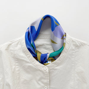 an abstract lotus print square silk scarf in blue, green, lilac and mustard yellow palette with hand-rolled hems tied as a classic neckerchief, paired with a light beige women's turtle neck shirt