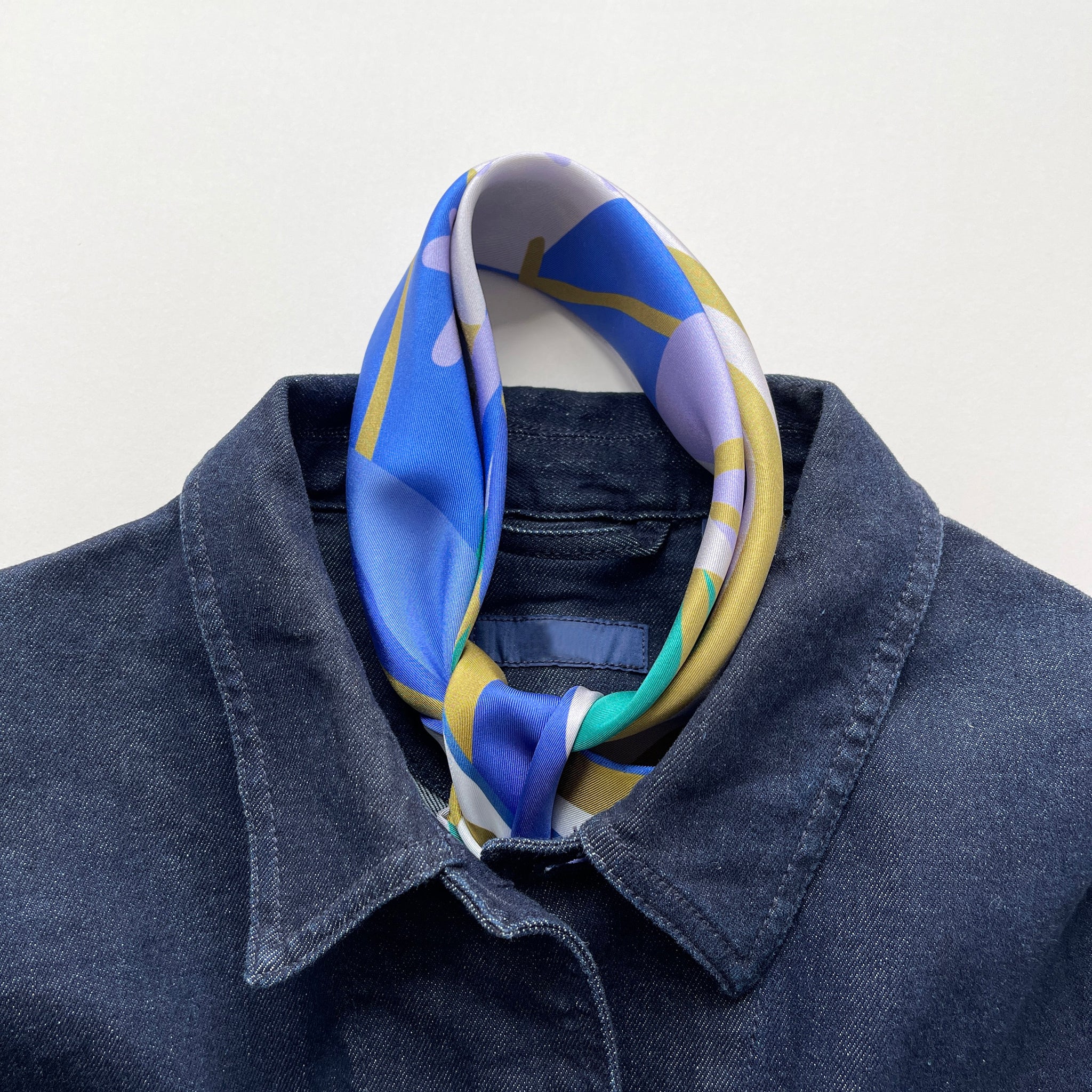 an abstract lotus print square silk scarf in blue, green, lilac and mustard yellow palette with hand-rolled hems tied as a classic neckerchief, paired with a men's denim jacket 