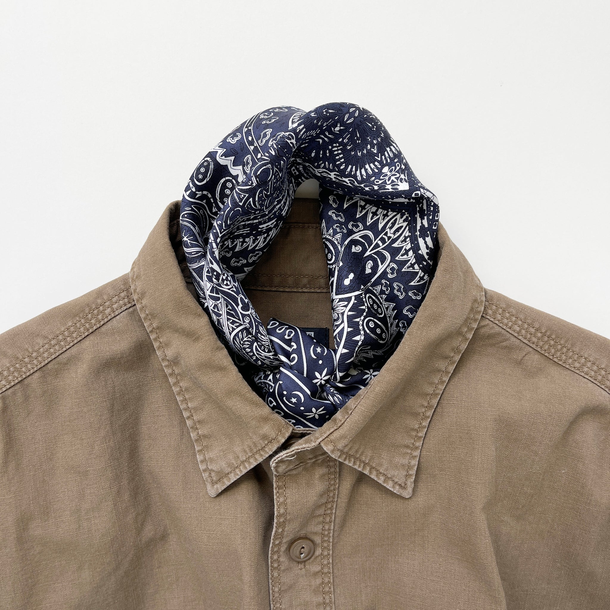 a navy blue mens neckerchief silk scarf featuring white and black symmetric pattern with striped hand-rolled edges, knotted as a neckerchief, paired with a khaki men's shirt
