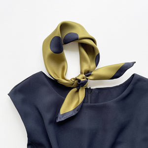 an olive green silk bandana scarf featuring black polka dots print, tied as a neckerchief, paired with a sleeveless black dress