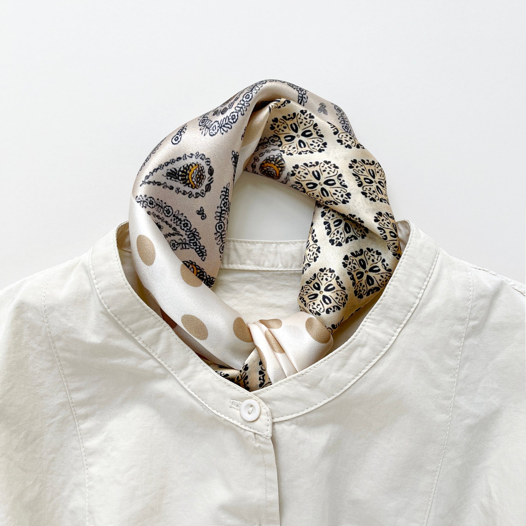 a beige and brown earth tones silk scarf featuring bohemian paisley and polka dot prints with brown edges, knotted as neck scarf, paired with a beige women's turtle neck shirt