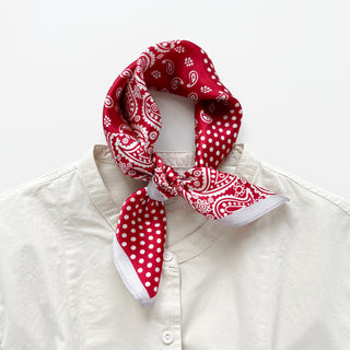 A 53x53cm red silk bandana scarf with white paisley and polka dot patterns, styled in a neat knot around the collar of a cream-colored button-up shirt. The scarf features alternating sections of intricate paisley designs and large polka dots, accented by a soft grey border. The luxurious silk fabric drapes elegantly, adding a stylish and polished touch to the casual outfit.