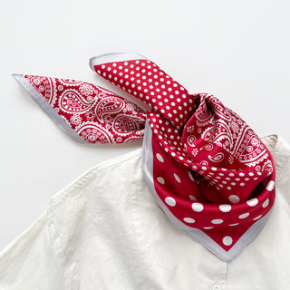  A 53x53cm red silk bandana scarf with white paisley and polka dot patterns, styled in a loose knot around the collar of a cream-colored button-up shirt. The scarf features alternating sections of intricate paisley designs and large polka dots, accented by a soft grey border. The luxurious silk fabric drapes elegantly, adding a stylish and polished touch to the casual outfit.