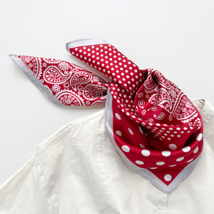 a vibrant red silk bandana scarf featuring paisley and polka dot pattern, knotted as a bandana bib, paired with a light beige turtle neck shirt