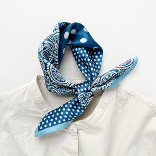 A 53x53cm blur silk bandana scarf with white paisley and polka dot patterns, styled in a neat knot around the collar of a cream-colored button-up shirt. The scarf features alternating sections of intricate paisley designs and large polka dots, accented by a lighter blue border. The luxurious silk fabric drapes elegantly, adding a stylish and polished touch to the casual outfit.