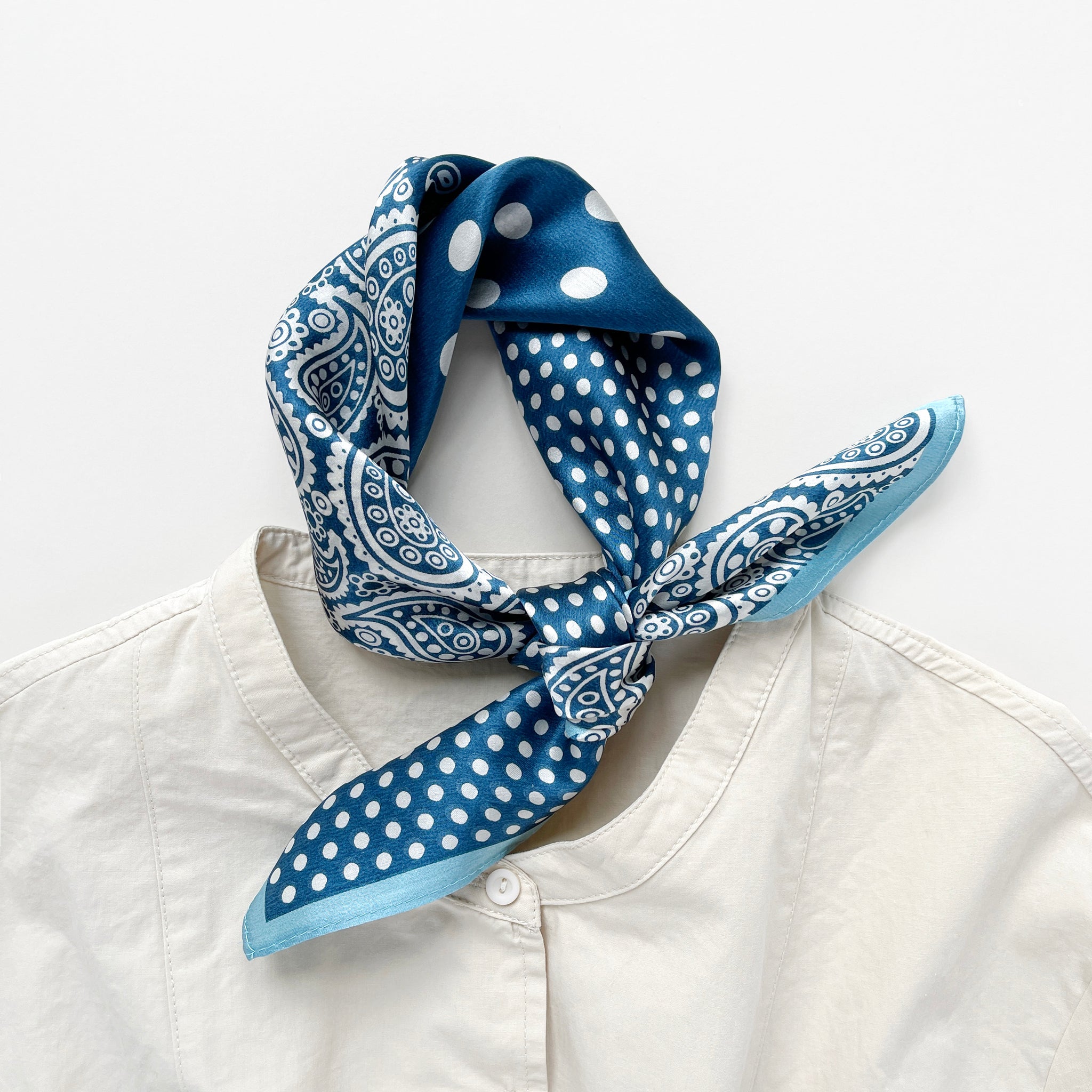 a vibrant blue silk bandana scarf featuring paisley and polka dot pattern, knotted as a neckerchief, paired with a light beige turtle neck shirt