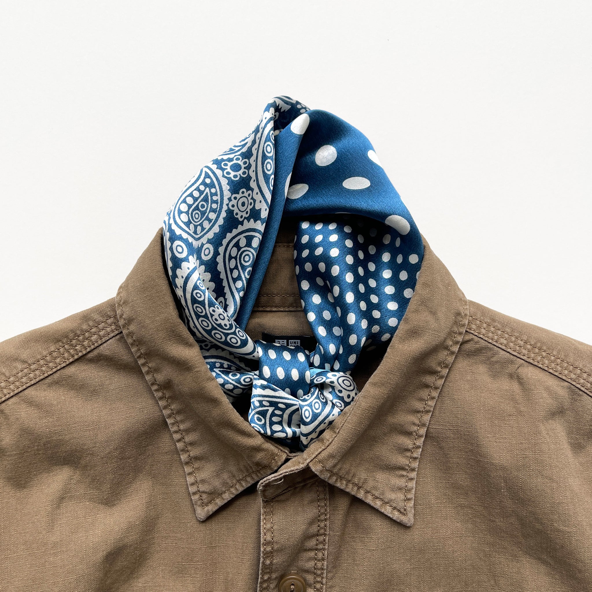a vibrant blue silk bandana scarf featuring paisley and polka dot pattern, knotted as a neckerchief, paired with a khaki men's shirt