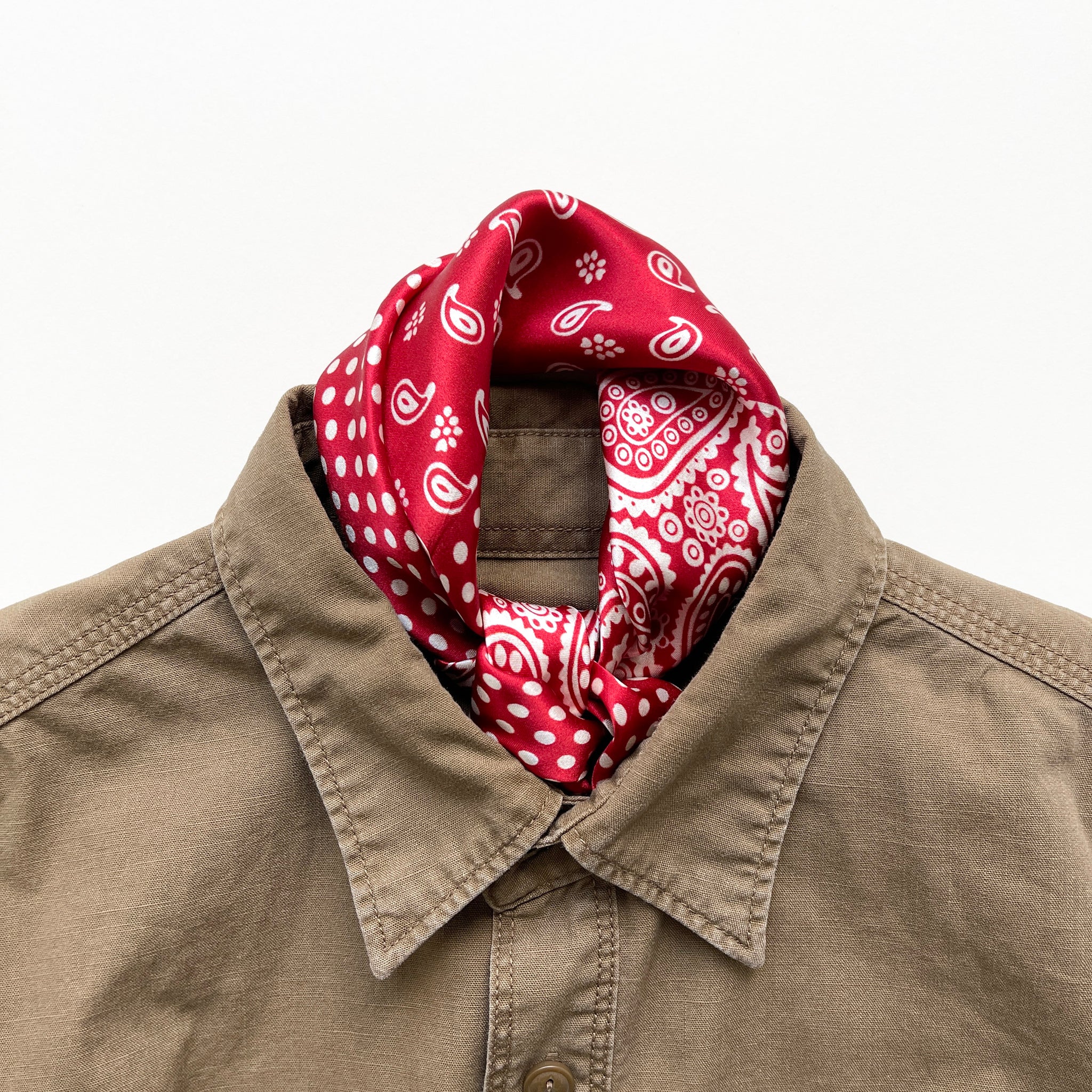 a vibrant red silk bandana scarf featuring paisley and polka dot pattern, knotted as a neckerchief, paired with a khaki men's shirt