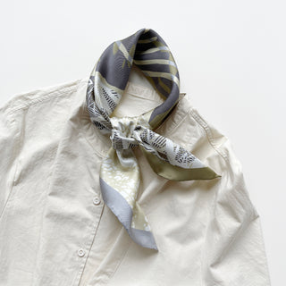 a forest leafy printed square silk scarf in sage green, grey and white tones with hand-rolled edges, tied as a classic neckerchief, paired with a light beige women's shirt